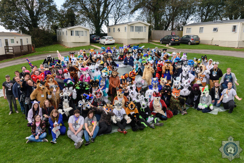 Furcation 2018 group photograph featuring both fursuiters and none taken on a small field with caravan park accommodation in the background.